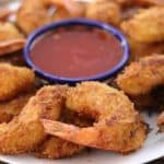 A plate of golden-brown fried shrimp arranged in a circle around a small bowl of red dipping sauce. The shrimp are crispy and have visible pieces of shredded coconut on the surface, indicating they are coconut shrimp. The bowl of sauce is placed at the center.