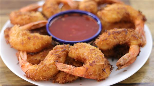 A plate of golden-brown fried shrimp arranged in a circle around a small bowl of red dipping sauce. The shrimp are crispy and have visible pieces of shredded coconut on the surface, indicating they are coconut shrimp. The bowl of sauce is placed at the center.