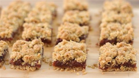 A close-up view of neatly arranged square date bars. Each bar has a crumbly oat topping and a dense, dark date filling in the middle, set on a wooden surface. The bars are aligned in multiple rows, creating a uniform pattern.