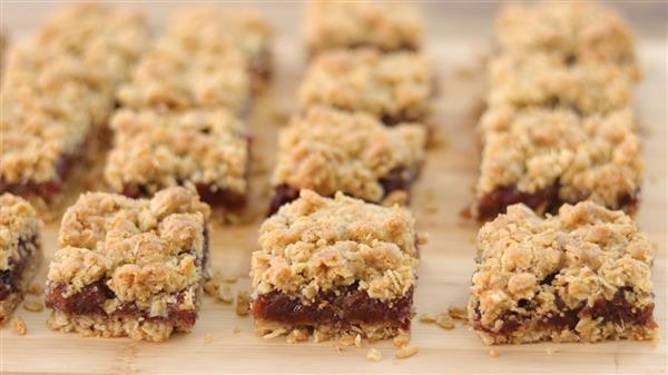 A close-up view of neatly arranged square date bars. Each bar has a crumbly oat topping and a dense, dark date filling in the middle, set on a wooden surface. The bars are aligned in multiple rows, creating a uniform pattern.