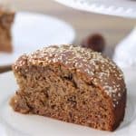 A slice of spiced date cake topped with sesame seeds, placed on a white plate. In the background, more slices of the cake are visible on a white cake stand and another white plate, partially out of focus. Some dates are also visible on the wooden surface.