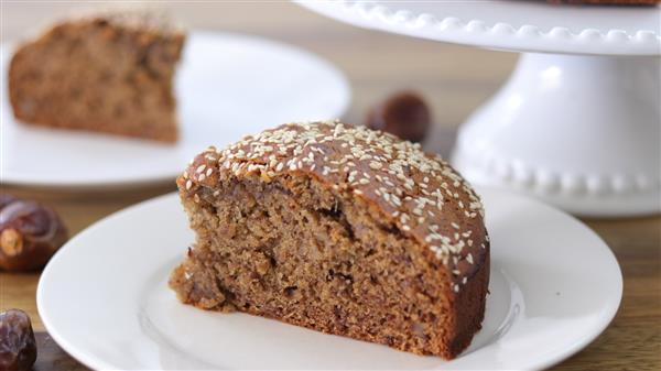 A slice of spiced date cake topped with sesame seeds, placed on a white plate. In the background, more slices of the cake are visible on a white cake stand and another white plate, partially out of focus. Some dates are also visible on the wooden surface.