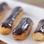 A row of chocolate eclairs is displayed on a rectangular white plate. The eclairs are golden brown and topped with a shiny layer of chocolate glaze. The plate is set on a wooden surface.