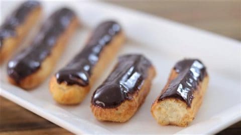 A row of chocolate eclairs is displayed on a rectangular white plate. The eclairs are golden brown and topped with a shiny layer of chocolate glaze. The plate is set on a wooden surface.