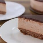 A slice of chocolate cheesecake with a dark chocolate glaze sits on a white plate. The cake has a crumbly chocolate crust and a smooth, creamy filling. In the background, another slice is on a separate plate, and the rest of the cheesecake is partially visible.