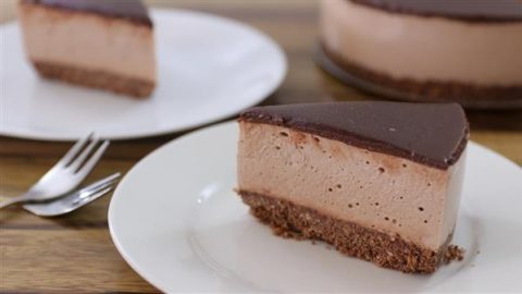 A slice of chocolate cheesecake with a dark chocolate glaze sits on a white plate. The cake has a crumbly chocolate crust and a smooth, creamy filling. In the background, another slice is on a separate plate, and the rest of the cheesecake is partially visible.