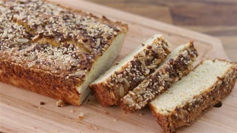 A loaf of bread topped with seeds and nuts, placed on a wooden cutting board. Two slices have been cut from the loaf, revealing a light, fluffy interior. The topping appears to include sunflower seeds, sesame seeds, and possibly other grains.