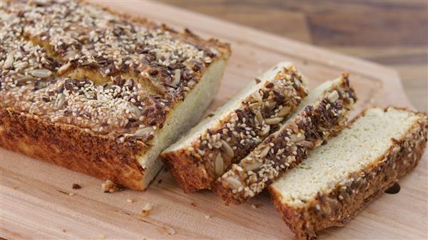 A loaf of bread topped with seeds and nuts, placed on a wooden cutting board. Two slices have been cut from the loaf, revealing a light, fluffy interior. The topping appears to include sunflower seeds, sesame seeds, and possibly other grains.