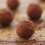 Close-up of multiple chocolate truffles covered in cocoa powder, arranged on a wooden surface. The truffles are scattered with slight cocoa powder dusting around them, giving a rustic and homemade appearance.