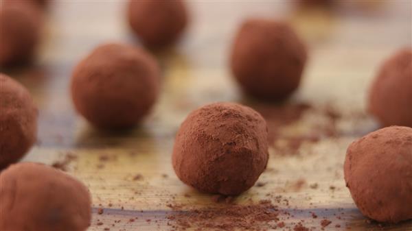 Close-up of multiple chocolate truffles covered in cocoa powder, arranged on a wooden surface. The truffles are scattered with slight cocoa powder dusting around them, giving a rustic and homemade appearance.