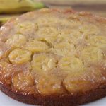 A freshly baked banana upside-down cake sits on a white plate. The cake features a glossy caramelized layer on top with banana slices arranged in a circular pattern. Two bananas rest on the wooden surface in the background for decoration.