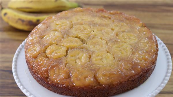 A freshly baked banana upside-down cake sits on a white plate. The cake features a glossy caramelized layer on top with banana slices arranged in a circular pattern. Two bananas rest on the wooden surface in the background for decoration.