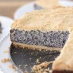 A round poppy seed cake with a crumbly golden crust is displayed on a black plate. A slice has been cut and removed, revealing the cake's interior. The background shows a wooden table with another plate featuring a slice of the same cake.