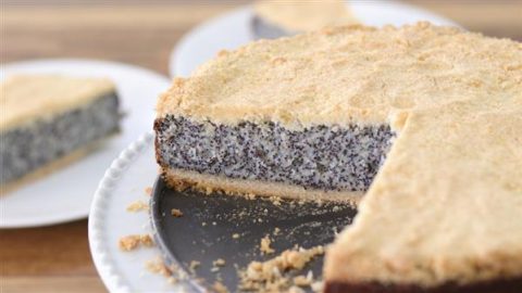 A round poppy seed cake with a crumbly golden crust is displayed on a black plate. A slice has been cut and removed, revealing the cake's interior. The background shows a wooden table with another plate featuring a slice of the same cake.