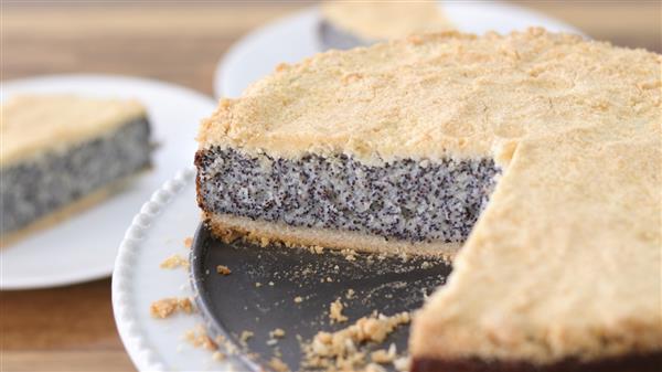 A round poppy seed cake with a crumbly golden crust is displayed on a black plate. A slice has been cut and removed, revealing the cake's interior. The background shows a wooden table with another plate featuring a slice of the same cake.