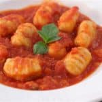 A white plate of gnocchi in tomato sauce is garnished with a sprig of fresh basil. The gnocchi are golden and slightly browned, nestled in a rich, vibrant red tomato sauce. The dish appears appetizing and ready to be served.