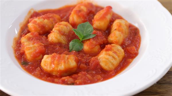 A white plate of gnocchi in tomato sauce is garnished with a sprig of fresh basil. The gnocchi are golden and slightly browned, nestled in a rich, vibrant red tomato sauce. The dish appears appetizing and ready to be served.