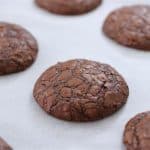 Chocolate cookies with a cracked surface are arranged on a white parchment-paper-lined baking sheet. The cookies appear freshly baked and have a rich, fudgy texture.