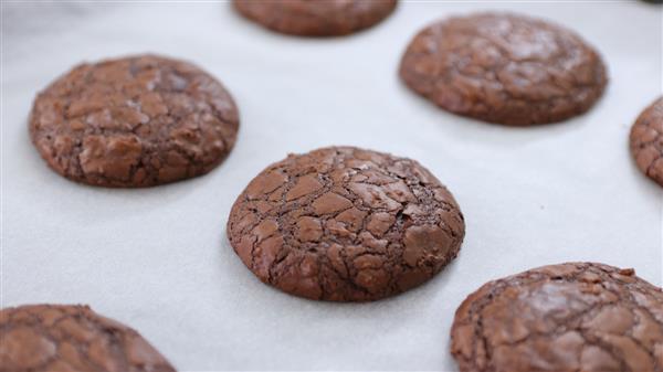 Chocolate cookies with a cracked surface are arranged on a white parchment-paper-lined baking sheet. The cookies appear freshly baked and have a rich, fudgy texture.