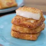 A stack of crispy, golden brown, pan-fried cheese slices sits on a light blue plate. A single thick slice of the cheese is placed neatly on top. A second plate of cheese slices is blurred in the background.
