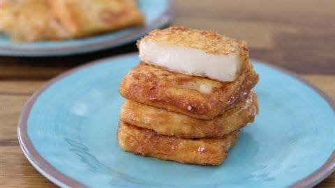 A stack of crispy, golden brown, pan-fried cheese slices sits on a light blue plate. A single thick slice of the cheese is placed neatly on top. A second plate of cheese slices is blurred in the background.