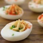 Close-up of deviled eggs on a wooden surface. The eggs are halved with creamy, piped yolk filling garnished with a sprinkle of paprika and chopped chives. The focus is on the front egg, with several more in the slightly blurred background.