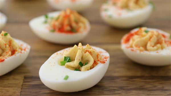 Close-up of deviled eggs on a wooden surface. The eggs are halved with creamy, piped yolk filling garnished with a sprinkle of paprika and chopped chives. The focus is on the front egg, with several more in the slightly blurred background.