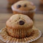 A close-up of several freshly baked chocolate chip muffins on a wooden surface. One muffin in the foreground is partially unwrapped from its paper liner, showcasing its moist, golden-brown texture and chocolate chip topping.