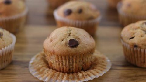 A close-up of several freshly baked chocolate chip muffins on a wooden surface. One muffin in the foreground is partially unwrapped from its paper liner, showcasing its moist, golden-brown texture and chocolate chip topping.