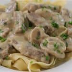 A plate of creamy beef stroganoff, featuring tender strips of beef and sliced mushrooms in a rich, creamy sauce, served over a bed of egg noodles and garnished with chopped green herbs.