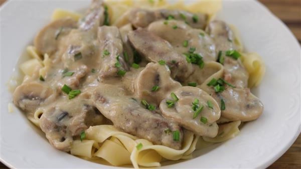 A plate of creamy beef stroganoff, featuring tender strips of beef and sliced mushrooms in a rich, creamy sauce, served over a bed of egg noodles and garnished with chopped green herbs.