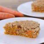 A slice of carrot cake is placed on a white plate with two whole carrots on a wooden table in the background. Another plate with another slice of carrot cake is partially visible behind it. The cake has a crumbly topping and visible carrot shreds.