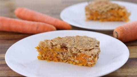 A slice of carrot cake is placed on a white plate with two whole carrots on a wooden table in the background. Another plate with another slice of carrot cake is partially visible behind it. The cake has a crumbly topping and visible carrot shreds.