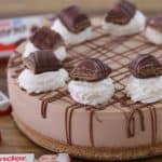 A creamy chocolate cheesecake topped with dollops of whipped cream and pieces of Kinder chocolate bars. The cheesecake features a graham cracker crust and is drizzled with chocolate. In the background, there are packages of Kinder chocolate bars.