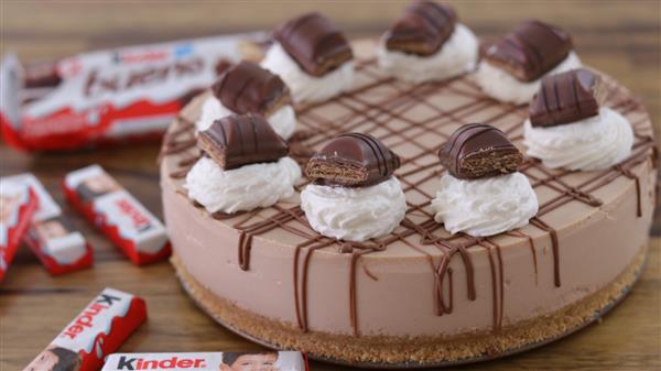 A creamy chocolate cheesecake topped with dollops of whipped cream and pieces of Kinder chocolate bars. The cheesecake features a graham cracker crust and is drizzled with chocolate. In the background, there are packages of Kinder chocolate bars.