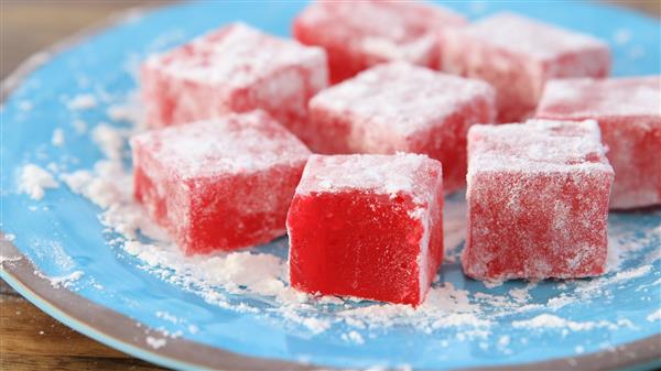 Close-up of several red Turkish delights dusted with powdered sugar, arranged on a blue plate. The cubes have a slightly translucent, chewy texture and are evenly coated with a fine layer of sugar, giving them a frosty appearance.