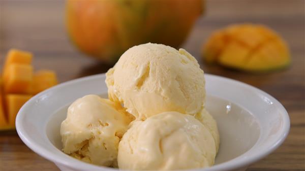 A white bowl contains three scoops of creamy yellow mango ice cream. In the background, there are pieces of cubed mango and a whole mango, slightly blurred. The setting appears to be on a wooden surface.