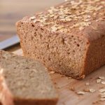 A freshly baked loaf of whole grain bread topped with oats is placed on a wooden cutting board. Two slices are cut from the loaf and rest in front of it. A bread knife lies nearby, and there are scattered oat flakes on the cutting board.