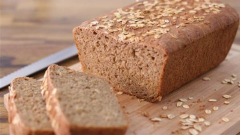 A freshly baked loaf of whole grain bread topped with oats is placed on a wooden cutting board. Two slices are cut from the loaf and rest in front of it. A bread knife lies nearby, and there are scattered oat flakes on the cutting board.