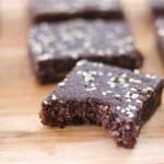 Homemade chocolate brownies with a sprinkled topping of white sesame seeds are arranged on a wooden surface. One brownie in the front row has a bite taken out of it, revealing a moist and dense texture inside.