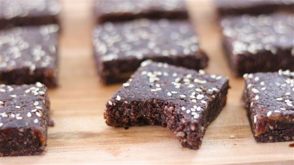 Homemade chocolate brownies with a sprinkled topping of white sesame seeds are arranged on a wooden surface. One brownie in the front row has a bite taken out of it, revealing a moist and dense texture inside.