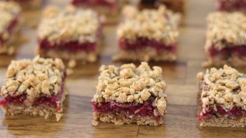 Close-up of several squares of dessert bars on a wooden surface. Each bar has a layer of granola-like crumble on top, a red filling in the middle, and an oat-looking base.