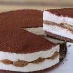 A close-up of a round tiramisu cake on a white plate. A slice has been lifted from the cake, showing layers of creamy mascarpone cheese, coffee-soaked ladyfingers, and a dusting of cocoa powder on top. The background is blurred.