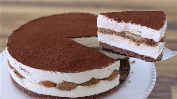 A close-up of a round tiramisu cake on a white plate. A slice has been lifted from the cake, showing layers of creamy mascarpone cheese, coffee-soaked ladyfingers, and a dusting of cocoa powder on top. The background is blurred.