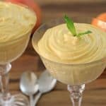 Two elegant glass cups contain creamy orange mousse, each adorned with a sprig of mint. The cups are placed on a wooden surface with two silver spoons beside them, and two whole oranges sit in the background.