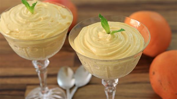 Two elegant glass cups contain creamy orange mousse, each adorned with a sprig of mint. The cups are placed on a wooden surface with two silver spoons beside them, and two whole oranges sit in the background.