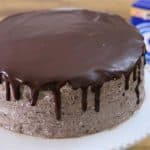 A round, Oreo-themed cake with smooth Oreo crumb frosting and a glossy chocolate ganache dripping down the sides, placed on a white cake stand. A blue package of Oreo cookies is visible in the background on a wooden surface.