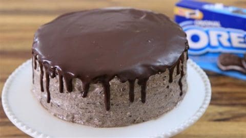 A round, Oreo-themed cake with smooth Oreo crumb frosting and a glossy chocolate ganache dripping down the sides, placed on a white cake stand. A blue package of Oreo cookies is visible in the background on a wooden surface.