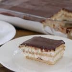 A slice of chocolate eclair cake sits on a white plate in the foreground. The cake features layers of graham crackers, creamy filling, and a chocolate topping. In the background, a glass baking dish holds the remaining eclair cake with one piece missing.