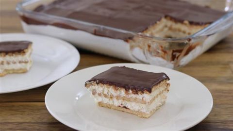 A slice of chocolate eclair cake sits on a white plate in the foreground. The cake features layers of graham crackers, creamy filling, and a chocolate topping. In the background, a glass baking dish holds the remaining eclair cake with one piece missing.